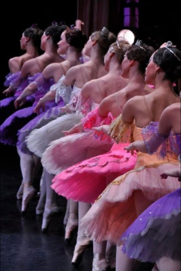 Eine Gruppe von Balletttänzerinnen in rosafarbenen Tutus sitzt auf einer Bühne, beleuchtet von einem Licht im Hintergrund, und bewegt sich in harmonischer Grazie.