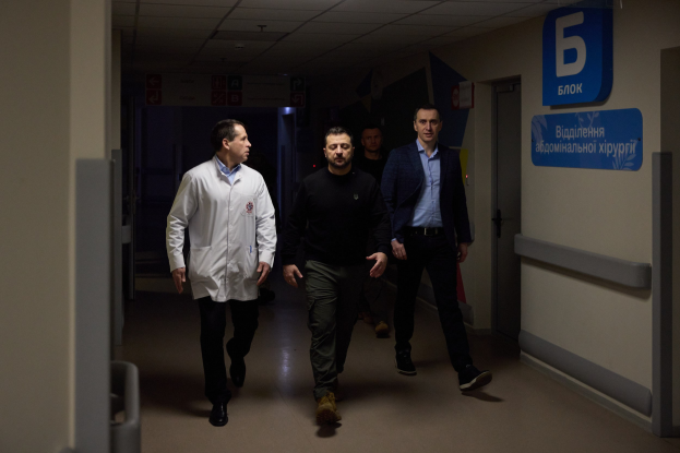 Eine Gruppe von Männern geht einen Krankenhausflur entlang mit Deckenleuchten, einer Tür an der rechten Wand und Texttafeln an der linken, die besorgt aussehen.