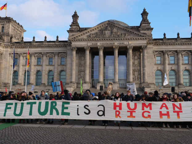 Gruppe von Menschen mit einem Banner 'Zukunft ist ein Menschenrecht' vor dem Reichstaggebäude in Berlin, Deutschland, mit sichtbaren Fenstern, Säulen und Bögen des Gebäudes, umgeben von Fahnenmasten, unter einem bewölkten Himmel.