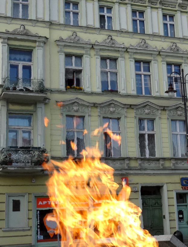 Ein großes Feuer brennt vor einem Gebäude mit Fenstern und Türen, einem Laternenmast und einer Tafel mit einer Aufschrift.