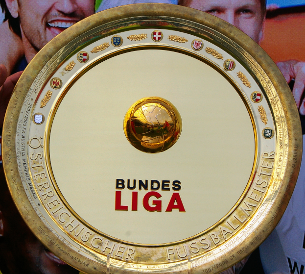 Ein Mann hält ein goldenes Schild mit der Aufschrift 'Bundesliga' hoch, im Hintergrund ist ein Banner mit drei Personen zu sehen.