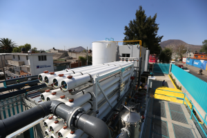 Eine große Gruppe von Rohren auf dem Dach eines Gebäudes, umgeben von Maschinen, Geländern, Fahrzeugen, Menschen, Gebäuden, Bäumen, Polen und Hügeln unter einem klaren blauen Himmel, wahrscheinlich im Zusammenhang mit der wegen Covid-19 erfolgten Schließung der mexikanischen Öl- und Gasindustrie.