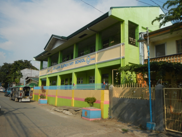 Schule mit grüner und gelber Fassade, umgeben von Fahrzeugen, Topfpflanzen, einem Zaun, einem Tor, einem Straßenpfahl, Bäumen, Drähten und einem bewölkten Himmel.