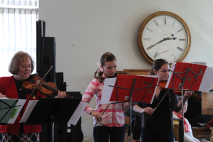 Drei Frauen auf der Bühne mit Violinen stehend und Musikständer vor sich, während ein sitzendes Publikum zuhört; eine Uhr hängt an der weißen Wand hinter ihnen, und ein weißer Vorhang ist links sichtbar.