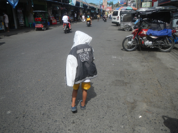 Ein junger Junge geht eine Straße voller Motorräder und anderer Fahrzeuge entlang, flankiert von Gebäuden mit Schildern, unter einem klaren blauen Himmel mit Bäumen und Polen im Hintergrund.