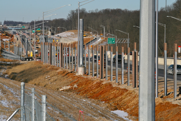Baustelle mit Fahrzeugen, Pfählen, Laternen, Schildern, einem Zaun, schneebedecktem Gras, Bäumen und einem Himmel im Hintergrund.