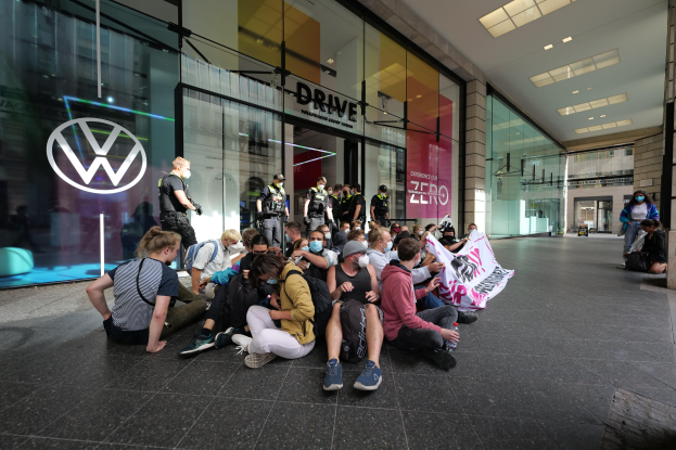 Gruppe von Menschen sitzt vor einem Volkswagen-Hndler und halt eine Plakette waehrend einer Demonstration.