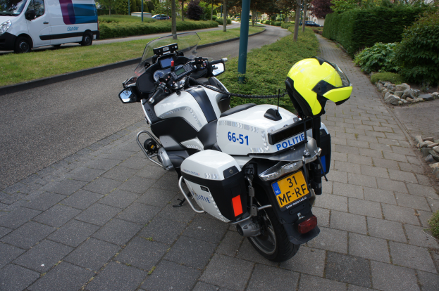 Ein polizeiliches Motorrad am Straßenrand geparkt mit einem Helm darauf, mit Bäumen, Pfählen, Gebäuden, Pflanzen, Gras, Steinen und anderen Fahrzeugen im Hintergrund.