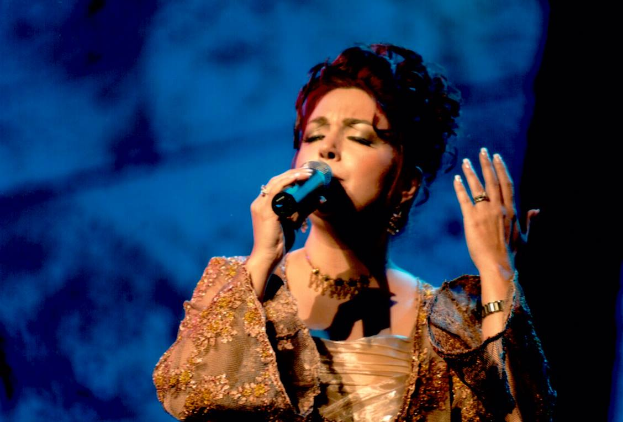 Eine Frau in einem goldenen Kleid singt in ein Mikrofon vor einem blauen und schwarzen Hintergrund, mit dem Text "Eurovision Song Contest" unten.