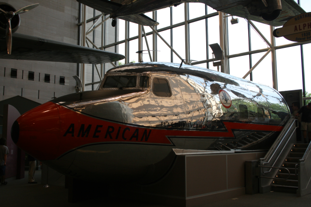 Ein Flugzeug von American Airlines in einem Museum ausgestellt, umgeben von Menschen auf dem Boden, Treppen auf der rechten Seite und ein Gebäude mit Fenstern, Bäumen und einem klaren blauen Himmel im Hintergrund.