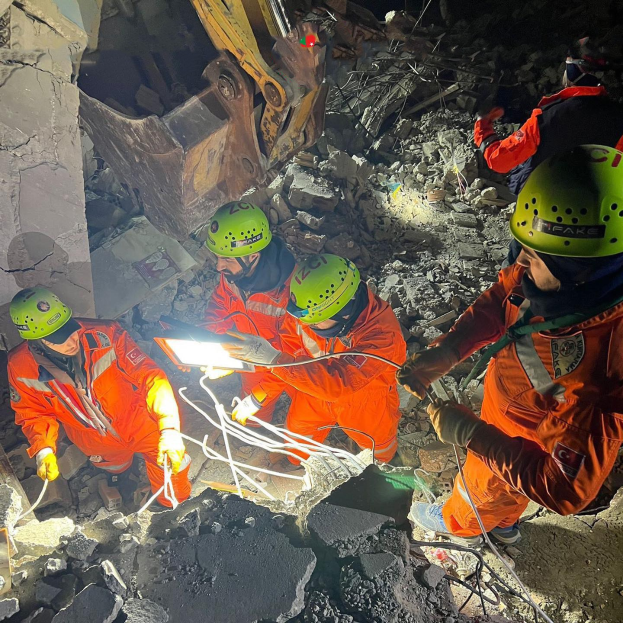 Eine Gruppe von Rettungskräften in orange Uniformen und grünen Helmen beseitigt Trümmmer von einem eingestürzten Gebäude, indem sie Werkzeuge verwenden, während im Hintergrund ein Kran arbeitet.
