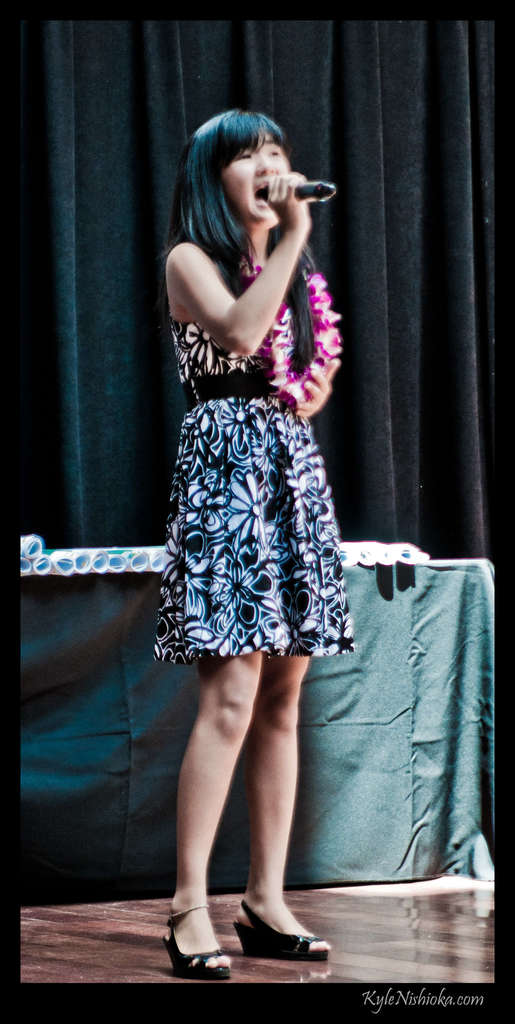 Eine Frau in einem bunten Kleid singt in ein Mikrofon auf der B├╝hne, mit einem Tisch mit Papieren und einem Vorhang im Hintergrund.