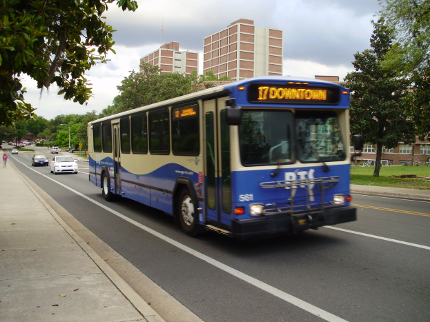 Ein blauer und weißer Shuttlebus fährt auf einer von hohen Gebäuden gesäumten Straße, mit einigen Passanten auf dem linken Gehweg und Bäumen, Masten und einem klaren blauen Himmel im Hintergrund.