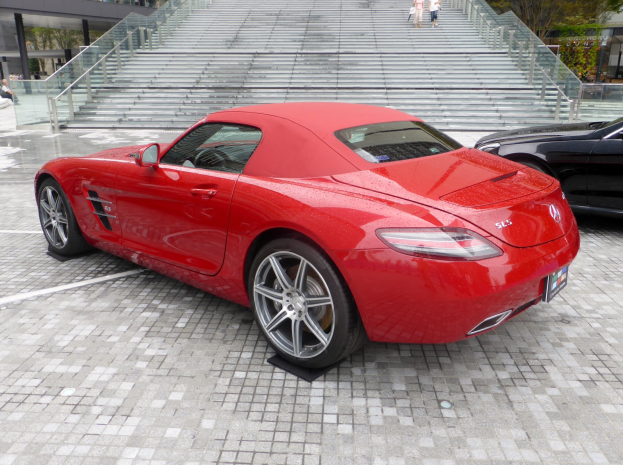 Ein roter Mercedes-Benz SLS AMG Roadster vor einem Gebäude geparkt, umgeben von Menschen, Treppen, Geländern, Bäumen und anderen Objekten.