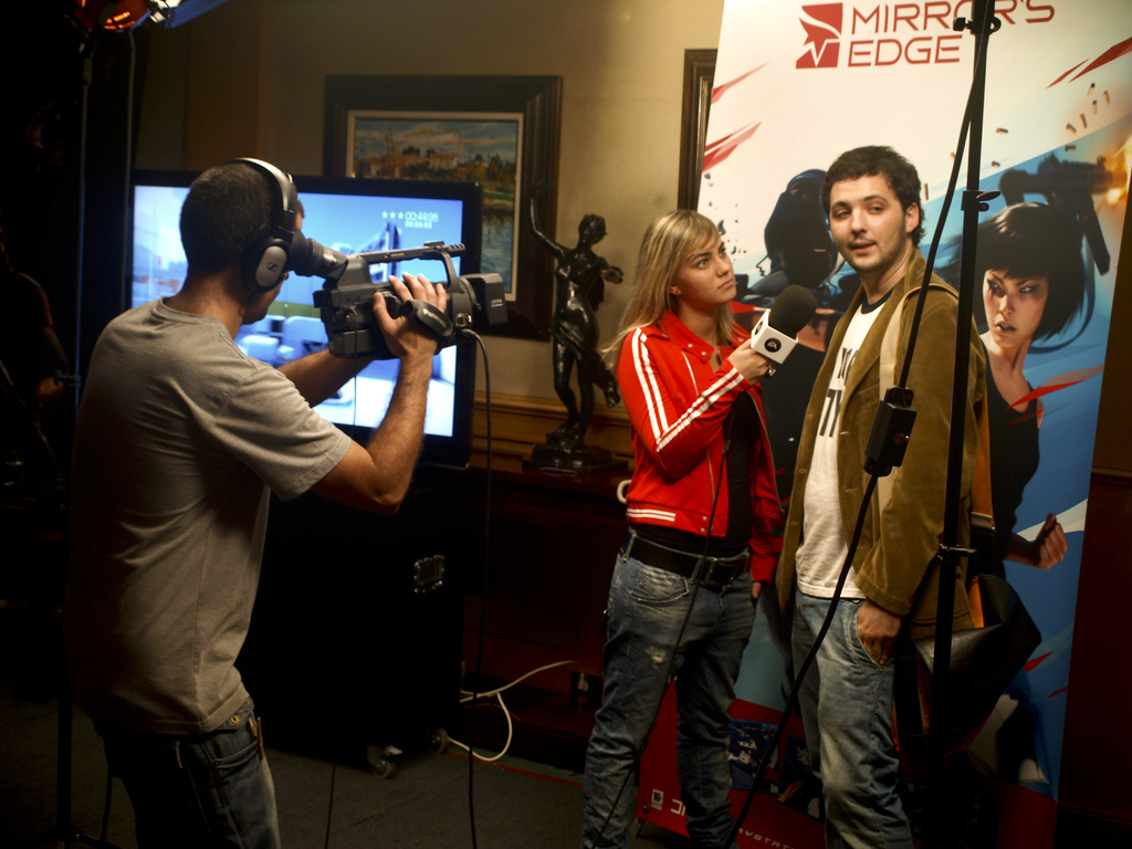 Ein Mann in einem Headset hält eine Videokamera und filmt ein Interview zwischen einem Mann und einer Frau, wobei im Hintergrund ein Fernseher und ein Foto Rahmen zu sehen sind.