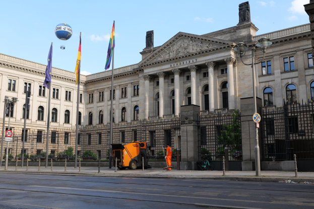 Großes modernes Gebäude mit vielen Fenstern, identifiziert als der Bundestag in Berlin, umgeben von Straßeninfrastruktur, Fußgängern, Flaggen und Bäumen unter einem bewölkten Himmel.