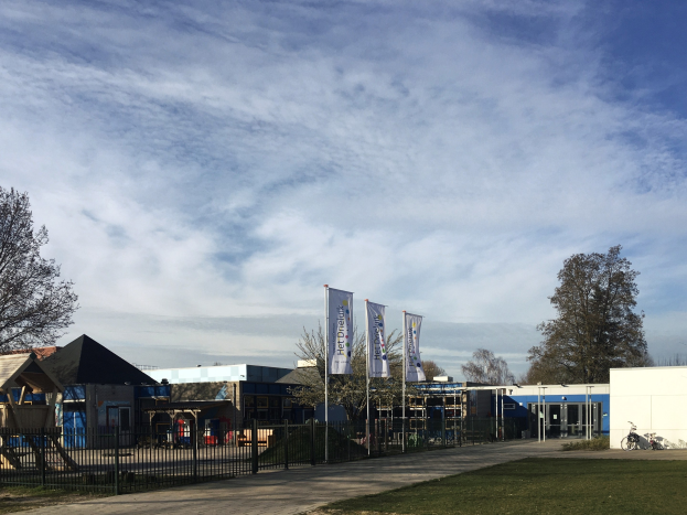 Großes Gebäude mit einem grasigen Bereich davor, umgeben von einem Metallzaun, mit einem Weg, Bäumen, Pfosten, Bannern, einem geparkten Fahrrad, nahen Häusern und einem bewölkten Himmel, als neuer Spielbereich für Kinder an der Schule identifiziert.