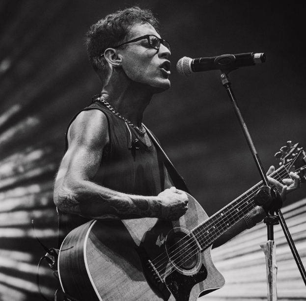 Ein Mann mit Brille spielt eine Gitarre vor einem Mikrofon, während er in der O2 Academy Brixton auftritt, dargestellt in Schwarzweiß.