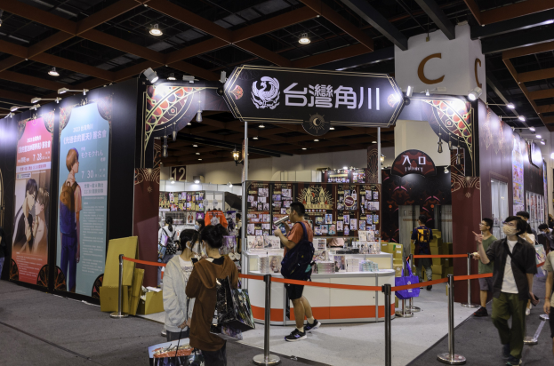Eine Gruppe von Menschen steht vor einem Messestand, einige halten Taschen, mit Bändern, Informationsplakaten, Deckenleuchten und Displayregalen im Hintergrund, wahrscheinlich für die Tokyo Anime Expo 2019.