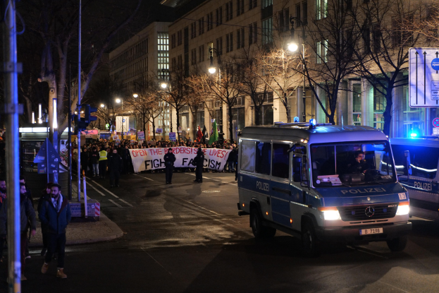 Eine Gruppe von Menschen geht nachts eine von Laternen beleuchtete Straße entlang an einem geparkten Polizeiwagen vorbei, mit Bäumen, Gebäuden und einer Fahne im Hintergrund.