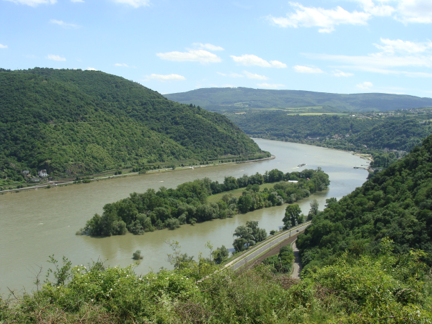 Luftaufnahme des sich durch ein grünes Tal schlängelnden Rheins mit einer Straße daneben und weißen Wolken am Himmel.