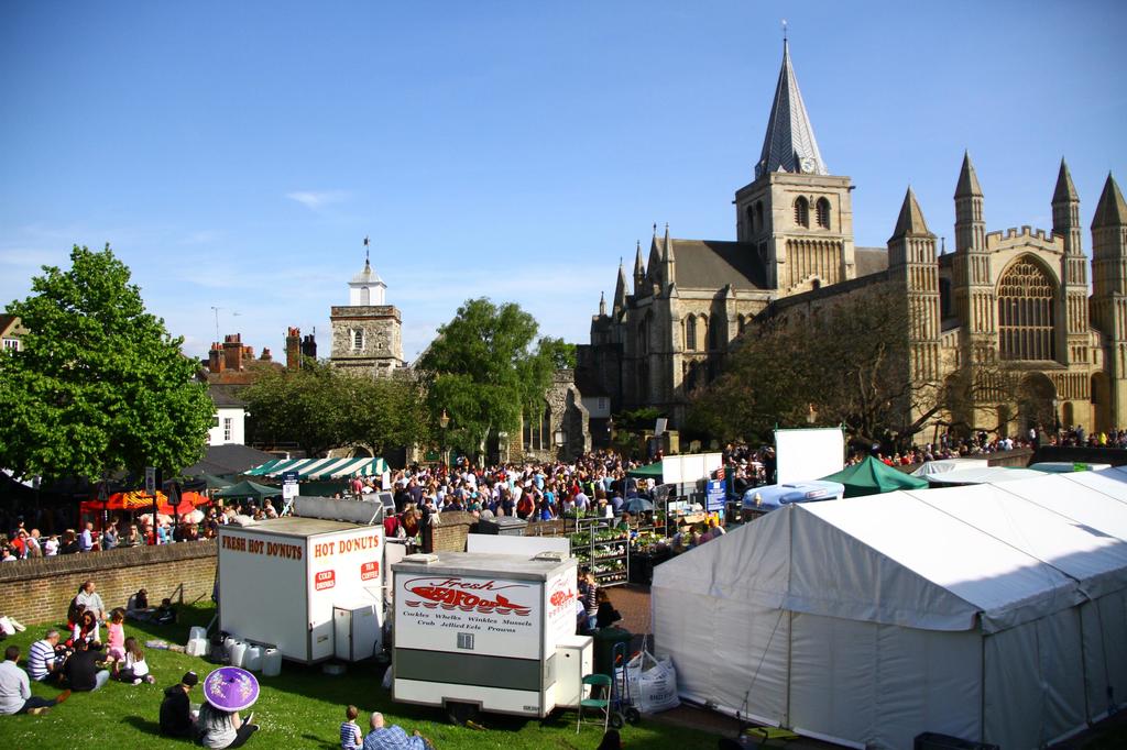 Historische Gebäude mit Bäumen davor, ein Turm im Hintergrund, Menschen auf einem Weg mit Geschäften und andere auf der Wiese in der Nähe.