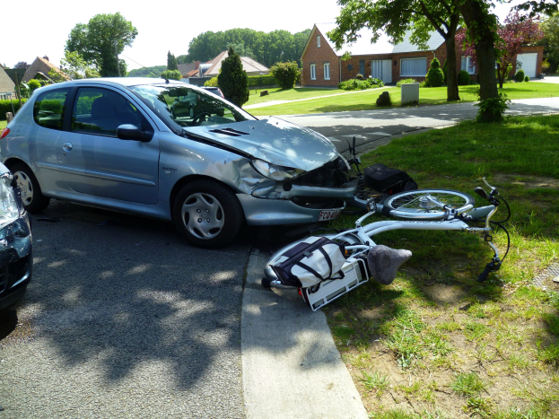 Ein Auto ist auf der Straße gegen ein Fahrrad geprallt, das auf dem Gras neben der Straße liegt, Häuser, Bäume und ein klarer blauer Himmel im Hintergrund.
