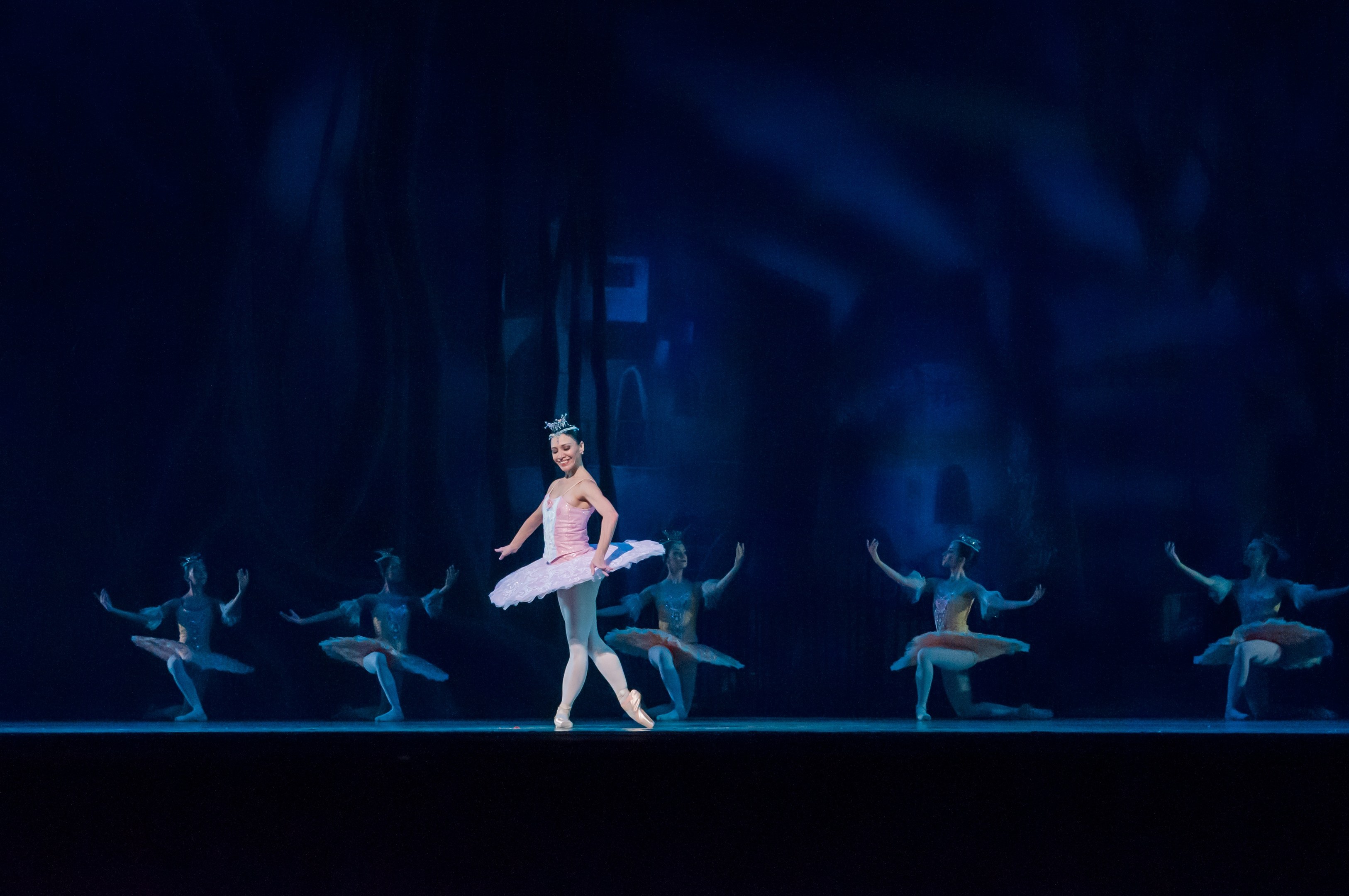 Eine Balletttänzerin in einem rosafarbenen Tutu und Spitzenschuhen tanzt auf einer Bühne mit anderen Tänzern in passenden Kostümen vor einem dunklen Hintergrund.