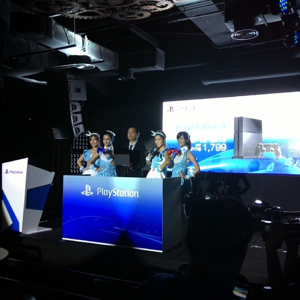 Menschen auf einer Bühne in der Nähe eines Podiums mit "Playstation"-Aufschrift, mit einem Projektionsschirm an der Wand dahinter.