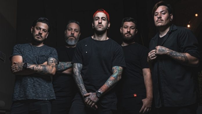 Eine Gruppe von Männern, die in einem dunklen Raum stehen, von rechts beleuchtet, während eines Interviews, das wahrscheinlich für Avenged Sevenfold stattfindet.