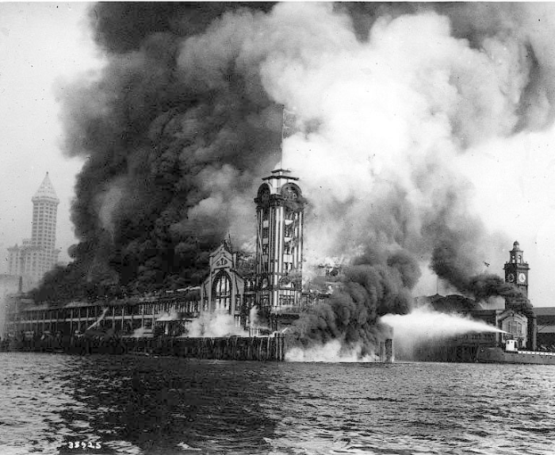 Schwarzes und weißes Foto eines großen, von Flammen eingehüllten Gebäudes mit Rauch, der daraus aufsteigt, einem Gewässer im Vordergrund und anderen Gebäuden sowie dem Himmel im Hintergrund.