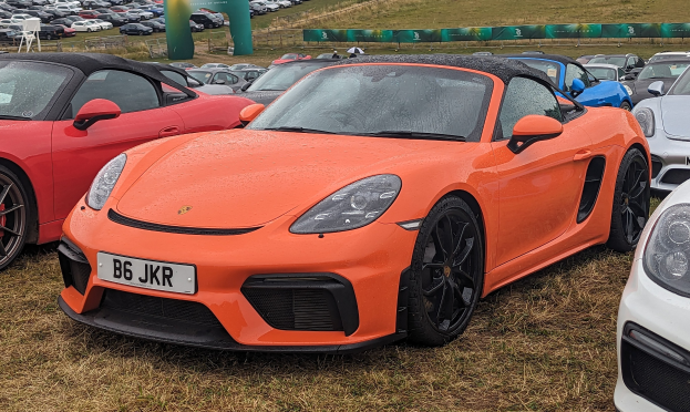 Roter Porsche 718 Boxster GTS beim Goodwood Festival of Speed mit Autos auf Gras, einem Banner, Bäumen und einem klaren blauen Himmel.