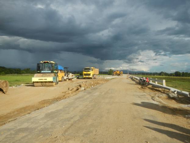 Baustelle mit Fahrzeugen, einem Zaun auf der rechten Seite, Gras, Bäumen im Hintergrund und einem bewölkten Himmel.