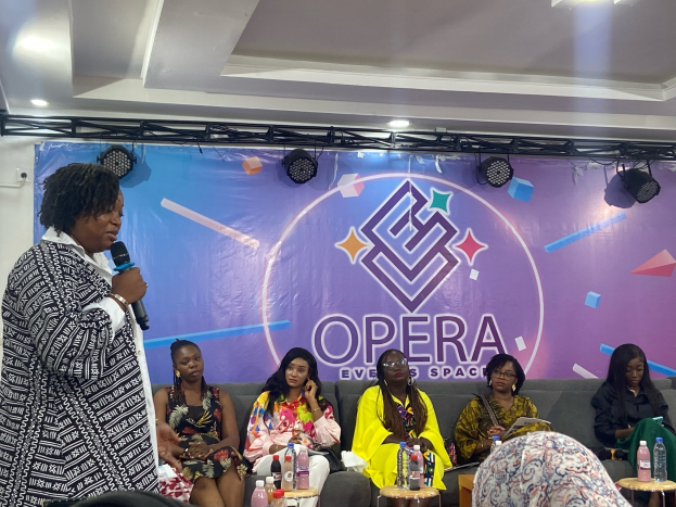 Frau mit Mikrofon vor sitzenden Frauen auf einer Couch, mit Flaschen und Gegenständen auf den Tischen im Vordergrund und einem Banner mit Text und Deckenbeleuchtung im Hintergrund.