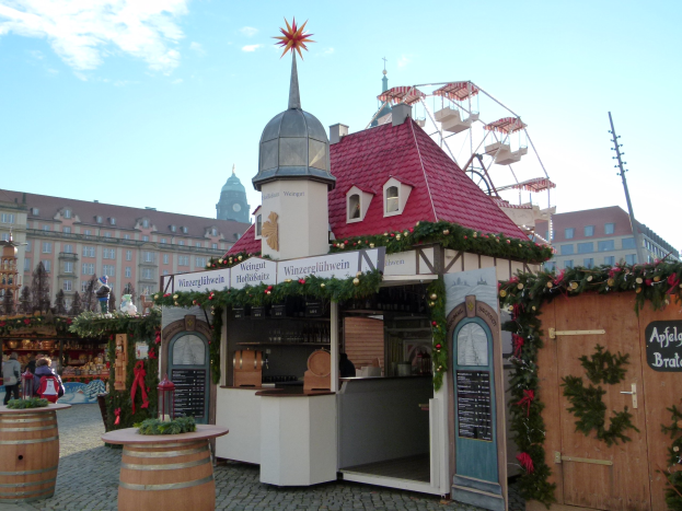 Kleines rotes Dachgebäude mit einem Riesenrad davor, umgeben von festlichen Oktoberfest-Dekorationen, Menschen und Gegenständen, vor einer Kulisse aus Gebäuden, Bäumen und einem bewölkten Himmel in München, Deutschland.