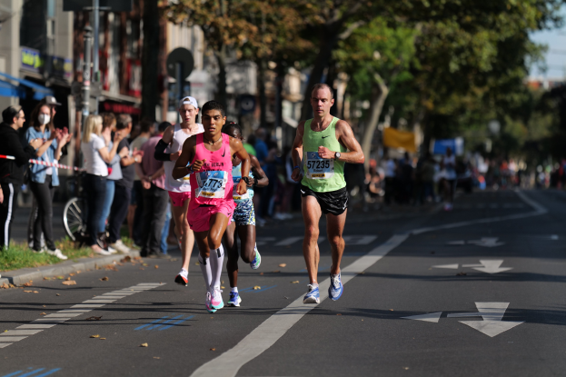 Gruppe von Menschen beim Marathon in einer Stadtstraße mit Zuschauern, Grünflächen und urbanen Elementen leicht unscharf im Hintergrund.