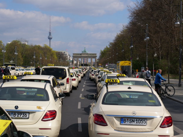 Eine lange Reihe von Taxis, die an einer belebten Straße in Berlin, Deutschland, geparkt sind, mit Fahrradfahrern und Fußgängern auf dem Gehweg, Laternenpfählen, Bäumen, Gebäuden, einem Bogen und einem Turm im Hintergrund bei einem bewölkten Himmel.