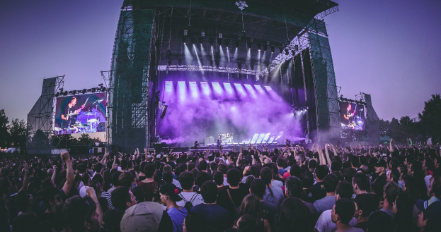 Eine große Menge Menschen bei einem Musikfestival schaut auf einer hell erleuchteten Bühne mit Instrumenten und Bildschirmen, die vor einem Gebäude, Bäumen und einem klaren blauen Himmel aufgebaut sind.