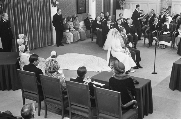 Schwarzes und weißes Foto einer Hochzeitszeremonie mit sitzenden und stehenden Menschen, einem Mann mit Mikrofon und einer Frau mit Blumenstrauß, einem Podium, zusätzlichen Mikrofonen, Blumenschmuck, einem Vorhang, einem gerahmten Foto an der Wand und einer Tür im Hintergrund.