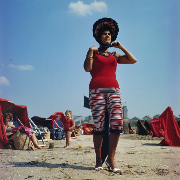 Frau in rotem Bikini auf einem Strand umgeben von sitzenden und stehenden Menschen, mit Zelten, Gebäuden und einer Flagge im Hintergrund.