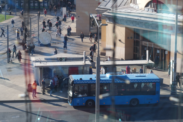 Ein blauer Bus fährt eine Straße mit hohen Gebäuden entlang, mit Fußgängern mit Taschen, Stangen und Grasflecken.