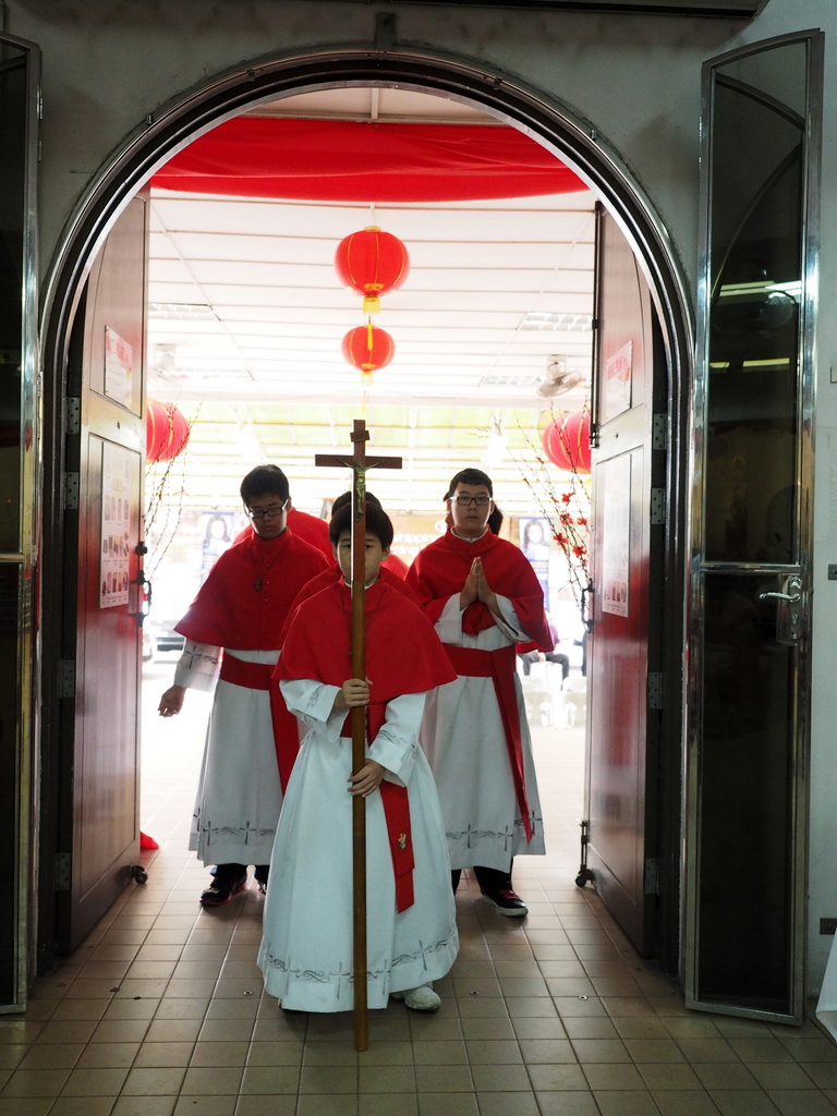 Menschen mit einem Kreuz in einer Kathedrale, mit Türen links und rechts und Blumen und dekorativen Elementen im Hintergrund.