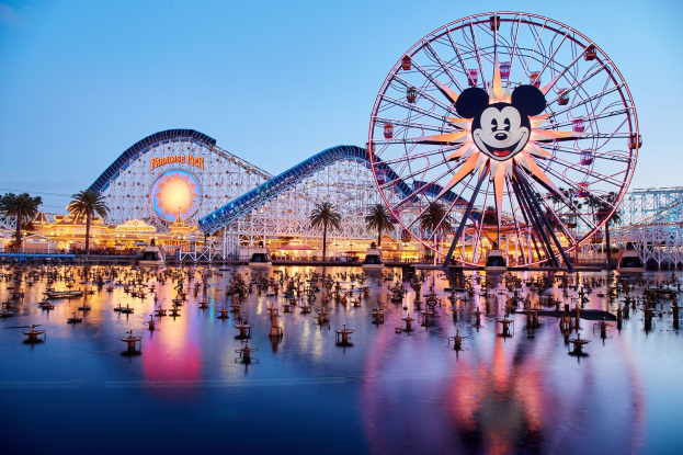 Eine lebendige Dämmerungsszene im Disneyland California Adventure mit einem riesigen Mickey-Maus-Rad im Vordergrund, einer Achterbahn im Hintergrund und Booten auf dem Wasser, mit einem Himmel aus Orangen, Pinks und Purpur und silhouettenhaften Bäumen.