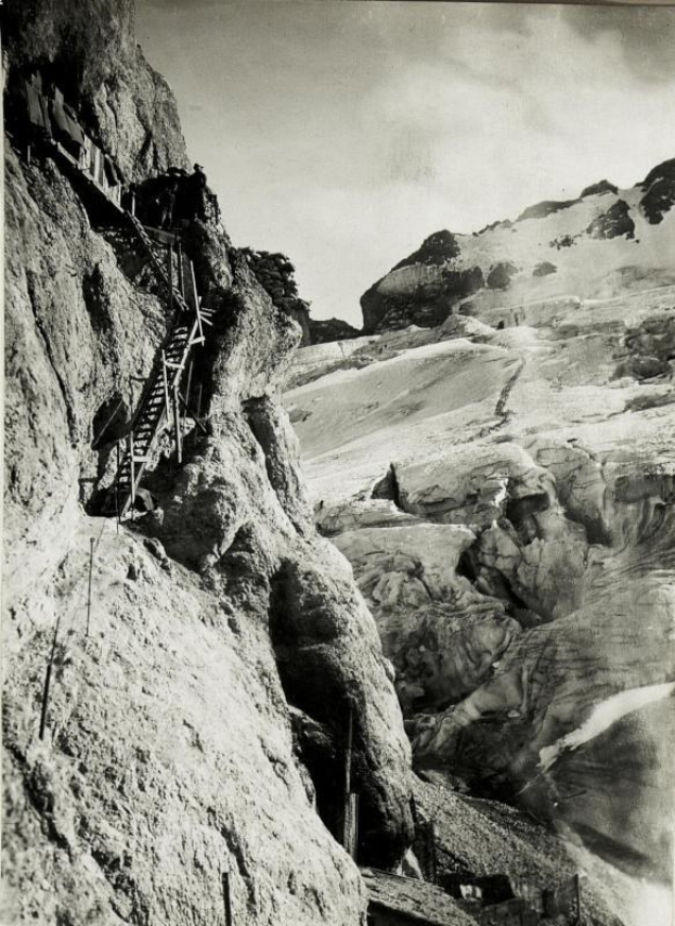 Schwarzes und weißes Foto von Kletterern, die einen schneebedeckten Berg hinaufsteigen, umgeben von anderen schneebedeckten Bergen und einem klaren Himmel, mit Text unten.