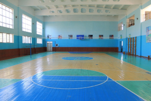 Großer Turnhalle mit blau-weißem Boden, Basketballfeld, Netz mit Stangen, Tür, Fenstern, Plakaten an den Wänden, Geländer und Dach.