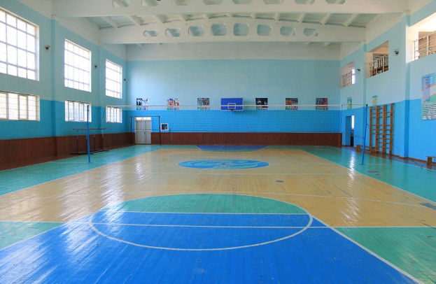 Großer Turnhalle mit blau-weißem Boden, Basketballfeld, Netz mit Stangen, Tür, Fenstern, Plakaten an den Wänden, Geländer und Dach.