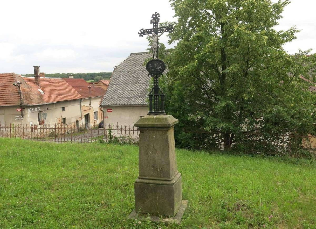 Ein großes Kreuz steht als Holocaust-Mahnmal in einer grünen Wiese, umgeben von einem Metallzaun, mit Bäumen, Häusern und einem bewölkten Himmel im Hintergrund.