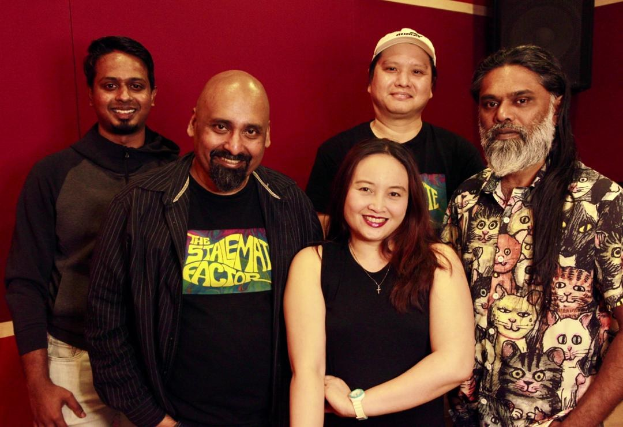 Eine Gruppe von Menschen, die zusammen in einem Aufnahmestudio posen und strahlend mit den Armen um die Schultern vor einer roten Wand stehen.