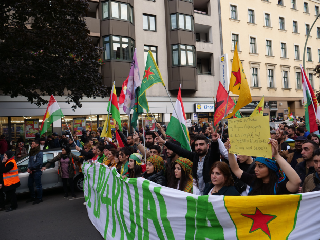 Eine große Gruppe von Menschen marschiert auf der Straße mit Fahnen und Transparenten, mit einem geparkten Auto und einem Baum auf der linken Seite und Gebäuden mit Fenstern und Namensschildern im Hintergrund, was auf eine algerische Demonstration hindeutet.
