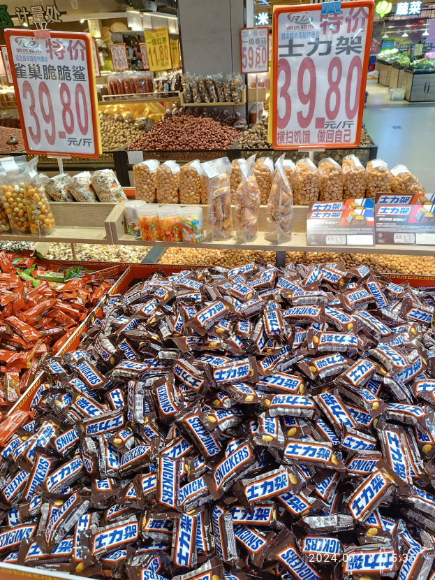 Vitrine mit verschiedenen Sorten von Süßigkeiten, einschließlich Snickers, in einem Geschäft mit Lebensmitteln auf Regalen und Hintergrunddetails.